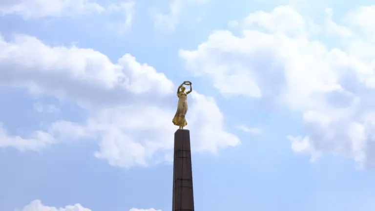 Złota figura Nike na wysokim postumencie na tle błękitnego nieba z białymi chmurami.