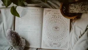 Otwarta książka z grafiką astrologiczną, kryształem i rośliną na miękkim tle