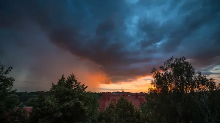 Słońce zachodzące nad drzewami i zabudowaniami w Szczecinie w pochmurny dzień.